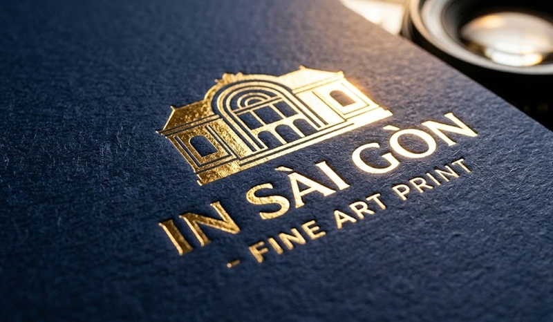 Cận cảnh logo ép kim nhũ vàng có độ lún sâu và phản quang như gương trên bề mặt giấy mỹ thuật.